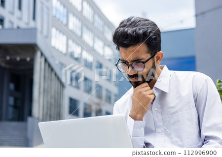 Close-up photo of a thoughtful and serious young Indian businessman in a white shirt sitting outside and staring blankly at a laptop screen. Close-up photo of a thoughtful and serious young Indian businessman in a white shirt sitting outside and staring blankly at a laptop screen. 112996900