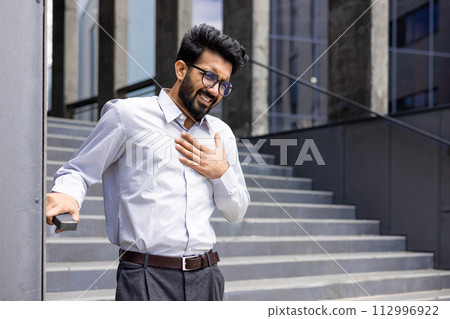 Contorted in pain, a young Indian male businessman stands by the wall of an office building and holds his hand over his heart. 112996922