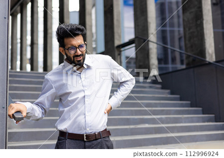Tired young Indian man in white shirt, office worker standing near building and holding hand behind back feeling severe lower back pain. Tired young Indian man in white shirt, office worker standing near building and holding hand behind back feeling severe lower back pain. 112996924