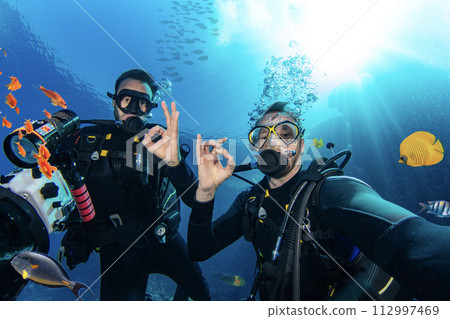 Scuba Divers Swimming in Deep Sea With Sunrays. Young Man DIver Eploring Sea Life. Scuba Divers Swimming in Deep Sea With Sunrays. Young Man DIver Eploring Sea Life. 112997469
