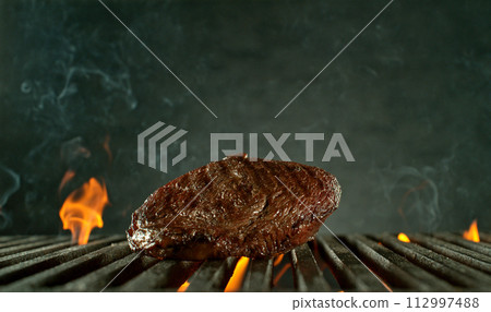 Close-up of tasty raw beef steak on cast-iron grate with fire flames 112997488