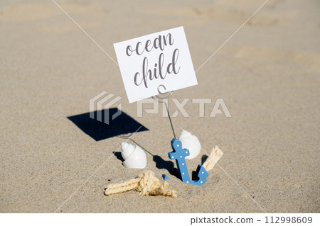 OCEAN CHILD text on paper greeting card in anchor paper holder and starfish seashell summer vacation decor. Sandy beach sun coast. Holiday concept postcard. Getting away Travel OCEAN CHILD text on paper greeting card in anchor paper holder and starfish seashell summer vacation decor. Sandy beach sun coast. Holiday concept postcard. Getting away Travel 112998609