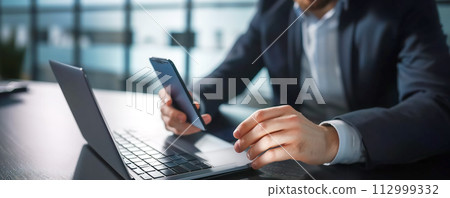 Man with smartphone in her hand working on the laptop in office. Place of work. Close-up view. Man with smartphone in her hand working on the laptop in office. Place of work. Close-up view. 112999332