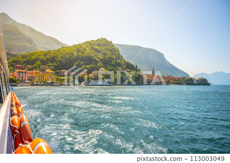 A boat is on the water with a view of a town in the background. The water is calm and the sky is clear. Como Lake, Italy A boat is on the water with a view of a town in the background. The water is calm and the sky is clear. Como Lake, Italy 113003049