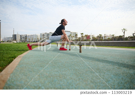 Determined sportsman in black t-shirt and gray sports shorts, warming up, doing lunges for stretching leg muscles. Determined sportsman in black t-shirt and gray sports shorts, warming up, doing lunges for stretching leg muscles. 113003219