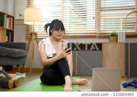 Young woman using smartphone on mat resting after fitness training at home Young woman using smartphone on mat resting after fitness training at home 113003463