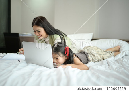 Cute little girl using laptop on the bed with her mother working in background Cute little girl using laptop on the bed with her mother working in background 113003519