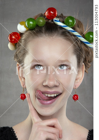 A funny teenager girl with a wreath of sweets on her head shows her tongue. A funny teenager girl with a wreath of sweets on her head shows her tongue. 113004793