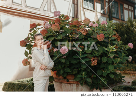 Attractive curly blonde woman walk on the city park street. Girl wear purple hoodie look happy and smiles. Woman make here me gesture standing near pink blooming bush flowers. Happy laughing girl. Attractive curly blonde woman walk on the city park street. Girl wear purple hoodie look happy and smiles. Woman make here me gesture standing near pink blooming bush flowers. Happy laughing girl. 113005271