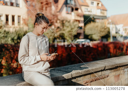 Young Fashionable Teenage Girl With Smartphone In Europian Park In Autumn Sitting At Smiling. Trendy Young Woman In Fall In Park Texting. Retouched, Vibrant Colors. Beautiful Blonde Teenage Girl 113005278