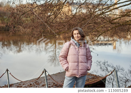 A young woman standing at the shore looking at the river in autumn sunny day. Street view, copy space for text, travel photo. Happy tourist woman on the bank of the river in autumn in warm clothes A young woman standing at the shore looking at the river in autumn sunny day. Street view, copy space for text, travel photo. Happy tourist woman on the bank of the river in autumn in warm clothes 113005289