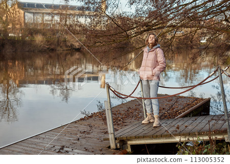 A young woman standing at the shore looking at the river in autumn sunny day. Street view, copy space for text, travel photo. Happy tourist woman on the bank of the river in autumn in warm clothes 113005352