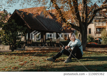 Professional Photographer Taking Picture Of Beautiful Autumn Park. Man Professional Photographer Sit With Camera And With Smartphone In Autumn Park. Retouched, Vibrant Colors, Brownish Tones. 113005362