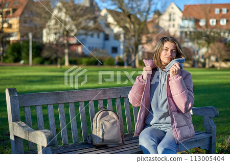 Winter Joy in Bitigheim-Bissingen: Beautiful Girl in Pink Jacket Sitting Amidst Half-Timbered Charm. beautiful girl in a pink winter jacket sitting on a bench in a park, set against the backdrop of 113005404
