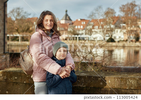 Riverside Family Harmony: Mother, 30 Years Old, and Son - Beautiful 8-Year-Old Boy, Standing by Neckar River and Historic Half-Timbered Town, Bietigheim-Bissingen, Germany, Autumn Riverside Family Harmony: Mother, 30 Years Old, and Son - Beautiful 8-Year-Old Boy, Standing by Neckar River and Historic Half-Timbered Town, Bietigheim-Bissingen, Germany, Autumn 113005424