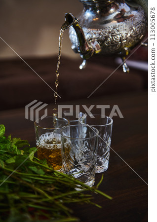 Pouring delicious Moroccan mint tea from silver tea pot into drinking glasses. bunch of fresh mint on the foreground Pouring delicious Moroccan mint tea from silver tea pot into drinking glasses. bunch of fresh mint on the foreground 113005678
