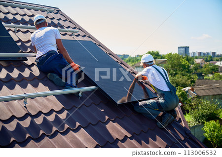 Workers building solar panel system on rooftop of house. Two men installers in helmets installing photovoltaic solar module outdoors. Alternative, green and renewable energy generation concept. 113006532