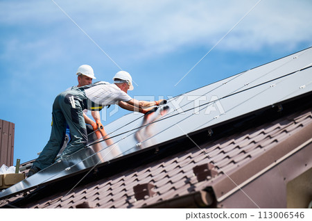 Workers building photovoltaic solar panel system on rooftop of house. Men technicians installing solar module with help of hex key outdoors. Alternative and renewable energy generation concept. 113006546