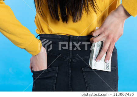 A young woman inserts a bundle of paper banknotes into the back pocket of jeans. 113007450