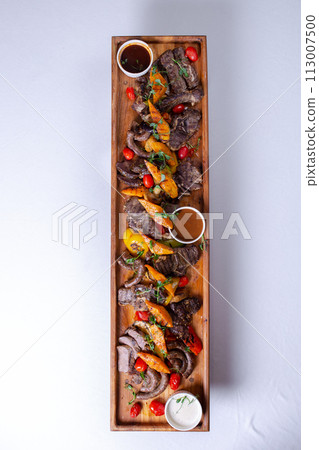 A long wooden platter filled with an assortment of meats and vegetables, including steak, sausage, chicken, potatoes, carrots, and peppers A long wooden platter filled with an assortment of meats and vegetables, including steak, sausage, chicken, potatoes, carrots, and peppers 113007500