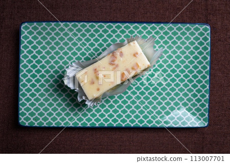 Square cake slice with icing and nuts on green plate, brown background Square cake slice with icing and nuts on green plate, brown background 113007701