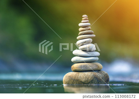 Close-up abstract image of wet rough natural brown uneven different sizes and forms stones balanced like pyramid pile landmark in shallow water on blurred blue-green misty copy space background. 113008172