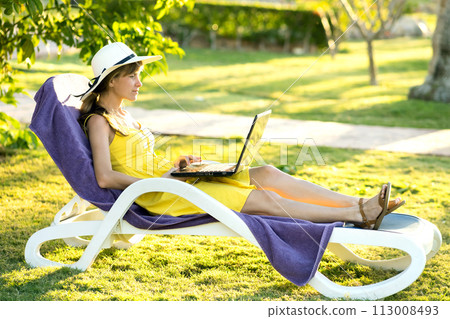 Young woman resting on chair on green grass lawn working on computer laptop connected to wireless internet in summer park. Doing business and studying remotely during quarantine on vacations concept. 113008493