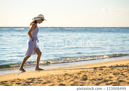 Young woman in straw hat and a dress walking alone on empty sand beach at sea shore. Lonely tourist girl looking at horizon over calm ocean surface on vacation trip. Young woman in straw hat and a dress walking alone on empty sand beach at sea shore. Lonely tourist girl looking at horizon over calm ocean surface on vacation trip. 113008499