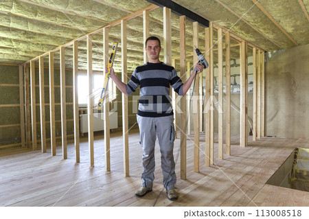 Young handsome worker with level and screwdriver looking in camera in big light attic room with oak floor, insulated ceiling and wooden frame for future walls. Construction and renovation concept. Young handsome worker with level and screwdriver looking in camera in big light attic room with oak floor, insulated ceiling and wooden frame for future walls. Construction and renovation concept. 113008518