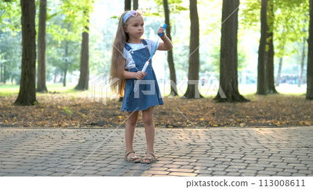 Little happy child girl blowing soap bubbles outside in green park. Outdoor summer activities concept. 113008611