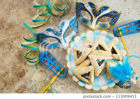 Jewish holiday Purim background with hamantaschen or hamans ears cookies, carnival mask and noisemaker Jewish holiday Purim background with hamantaschen or hamans ears cookies, carnival mask and noisemaker 113009451