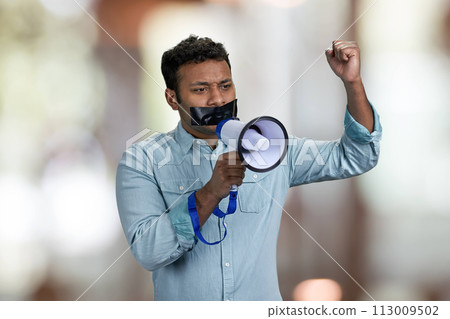 Young man acivist with taped mouth trying to speak into megaphone. Abstract bokeh background. 113009502