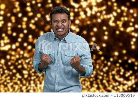 Angry aggressive guy grimacing and shaking fists. Festive bokeh background. Human negative expressions. Angry aggressive guy grimacing and shaking fists. Festive bokeh background. Human negative expressions. 113009519