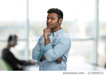 Thoughtful young man holding hand under chin. Office interior blur background. Young man with suspicious expression. Thoughtful young man holding hand under chin. Office interior blur background. Young man with suspicious expression. 113009523