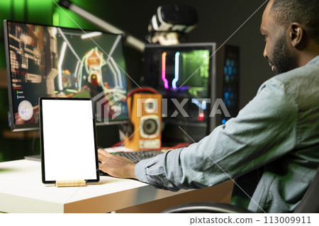 Mockup tablet on desk next to BIPOC man playing SF videogame on computer, talking into headset with friends. Isolated screen portable device and gamer using headphones to chat with team in game 113009911
