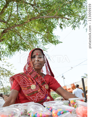Elderly woman in red saree selling food and sweets at street market in Jaipur 113009939