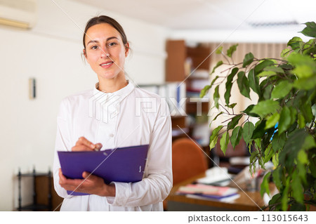 Portrait of a young female manager standing in a office and making notes Portrait of a young female manager standing in a office and making notes 113015643
