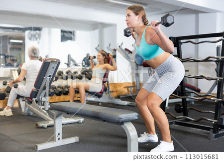 Slender girl trains muscles of arms and back using barbell and differentiated weighting in gym 113015681