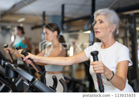 Slender athletic mature woman during cardio exercises on elliptical simulator in fitness center 113015835