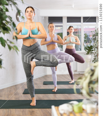 Girl exercising with group at yoga studio, standing in warrior one leg pose on fitness mat Girl exercising with group at yoga studio, standing in warrior one leg pose on fitness mat 113015890