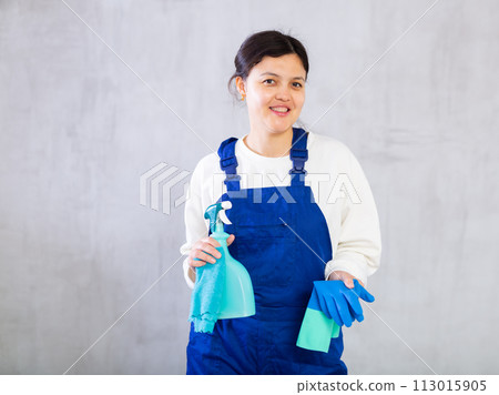 Smiling young cleaning lady in blue overalls with rag and detergent Smiling young cleaning lady in blue overalls with rag and detergent 113015905