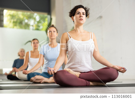 Serious young Latin female sitting in Lotus Pose during group yoga class in fitness studio Serious young Latin female sitting in Lotus Pose during group yoga class in fitness studio 113016001