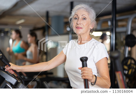 Slender athletic mature woman during cardio exercises on elliptical simulator in fitness center Slender athletic mature woman during cardio exercises on elliptical simulator in fitness center 113016004