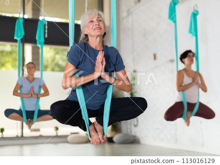 Old European female balancing in Lotus Pose Padmasana in blue hammock during fly yoga in fitness club 113016129