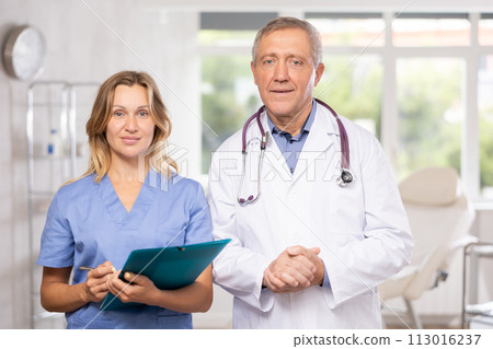 Elderly male doctor with female assistant standing in medical office 113016237