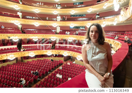 Interested young adult brunette sitting on balcony of classic concert hall Interested young adult brunette sitting on balcony of classic concert hall 113016349
