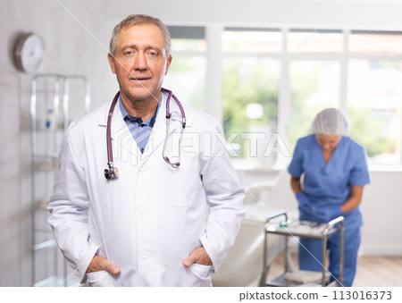Old man doctor posing in doctor's cabinet with clinical chair 113016373
