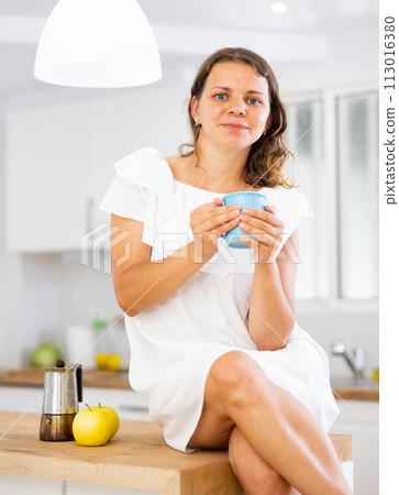 Portrait of young woman in white dress drinking morning coffee in modern kitchen 113016380