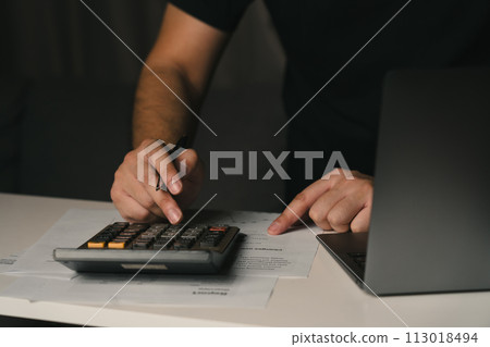 Asian man is using a calculator to calculate his family's monthly miscellaneous expenses at home and record them in his laptop computer. Asian man is using a calculator to calculate his family's monthly miscellaneous expenses at home and record them in his laptop computer. 113018494