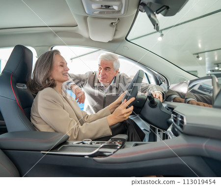 An elderly couple chooses a new car at a car dealership. Mature woman driving. An elderly couple chooses a new car at a car dealership. Mature woman driving. 113019454
